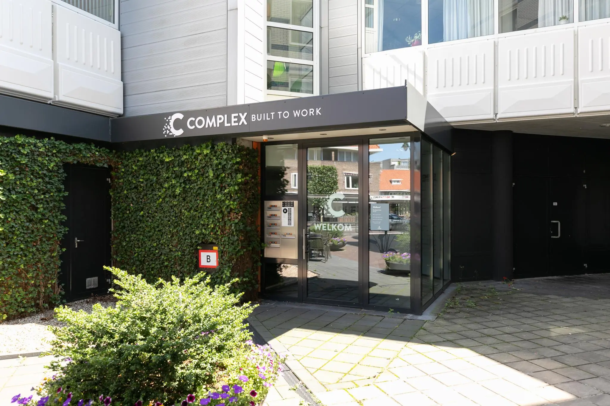 Ingang van het bedrijfsverzamelgebouw Complex aan de 1e Hogeweg met glasdeur, begroeiing en een B-ingangsaanduiding.